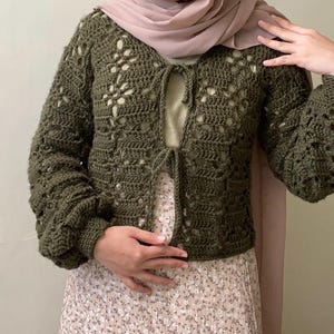Pu&ograve; includere: Un cardigan di uncinetto verde con un davanti a cravatta e maniche lunghe. Il cardigan presenta un delicato motivo traforato.