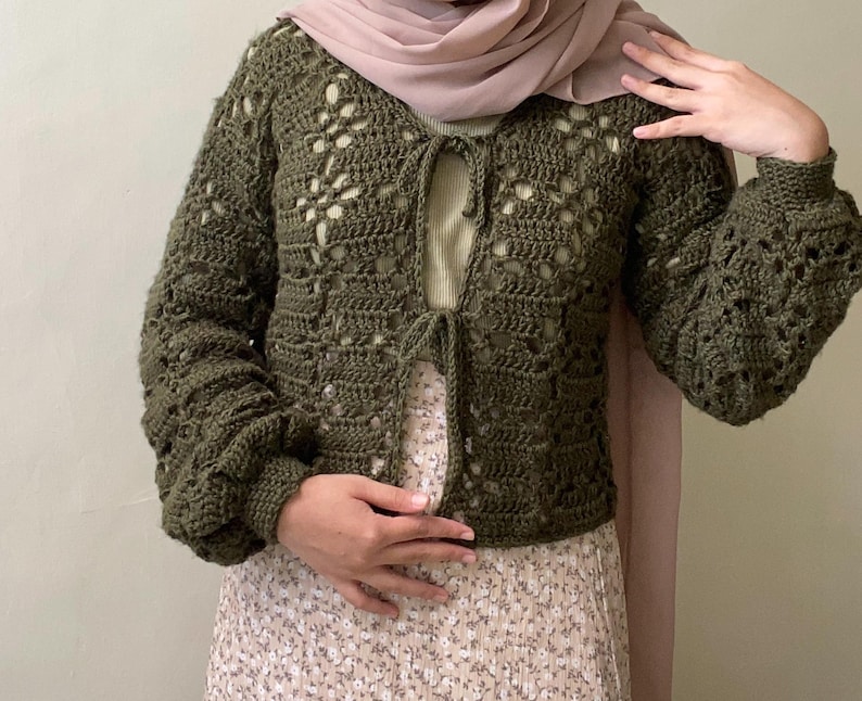 Pu&ograve; includere: Un cardigan di uncinetto verde con un davanti a cravatta e maniche lunghe. Il cardigan presenta un delicato motivo traforato.