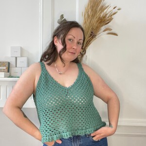 Op de afbeelding: Een groene gehaakte tanktop met een v-hals en een gegolfde zoom. De tanktop wordt gedragen over een blauwe jeans.