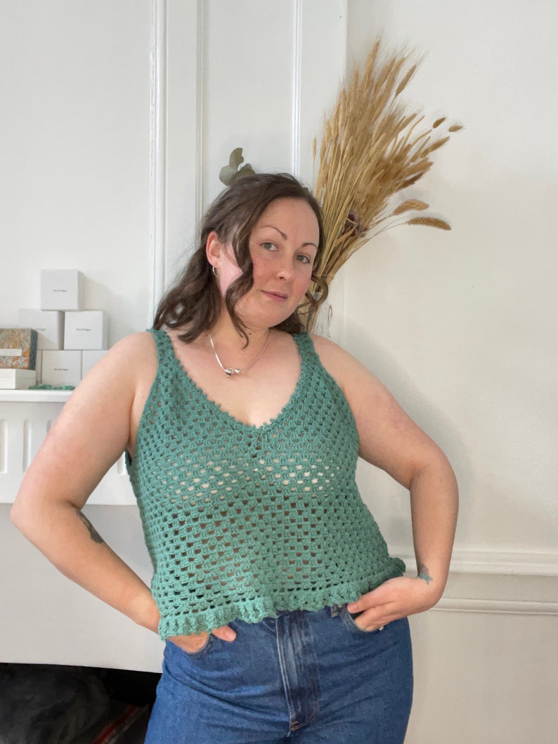 Op de afbeelding: Een groene gehaakte tanktop met een v-hals en een gegolfde zoom. De tanktop wordt gedragen over een blauwe jeans.