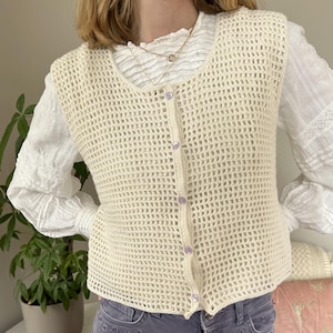 Op de afbeelding: Een cr&egrave;mekleurige gehaakte vest met een knoopsluiting aan de voorkant. De vest heeft een losse, open structuur en is mouwloos.