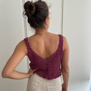 Op de afbeelding: Een close-up van een vrouw die een bordeauxrode gehaakte tanktop draagt met een diepe V-hals. De top heeft een delicaat gehaakt patroon en een gerimpelde zoom.