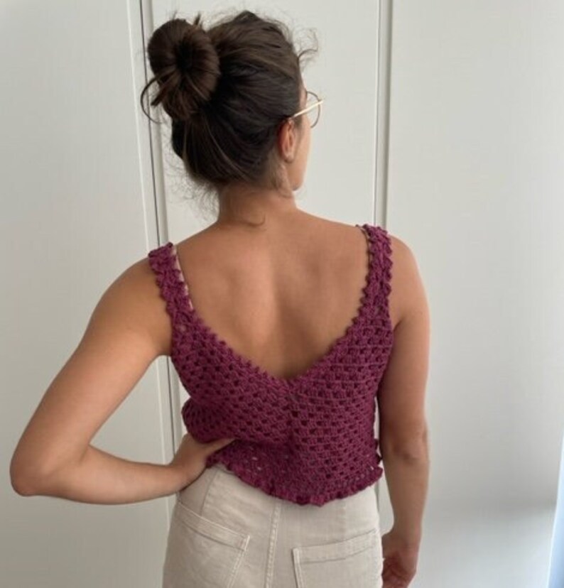 Op de afbeelding: Een close-up van een vrouw die een bordeauxrode gehaakte tanktop draagt met een diepe V-hals. De top heeft een delicaat gehaakt patroon en een gerimpelde zoom.