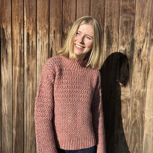 May include: A woman wearing a pink knitted sweater with a textured pattern. The sweater has long sleeves and a slightly cropped length. She is standing in front of a wooden wall.