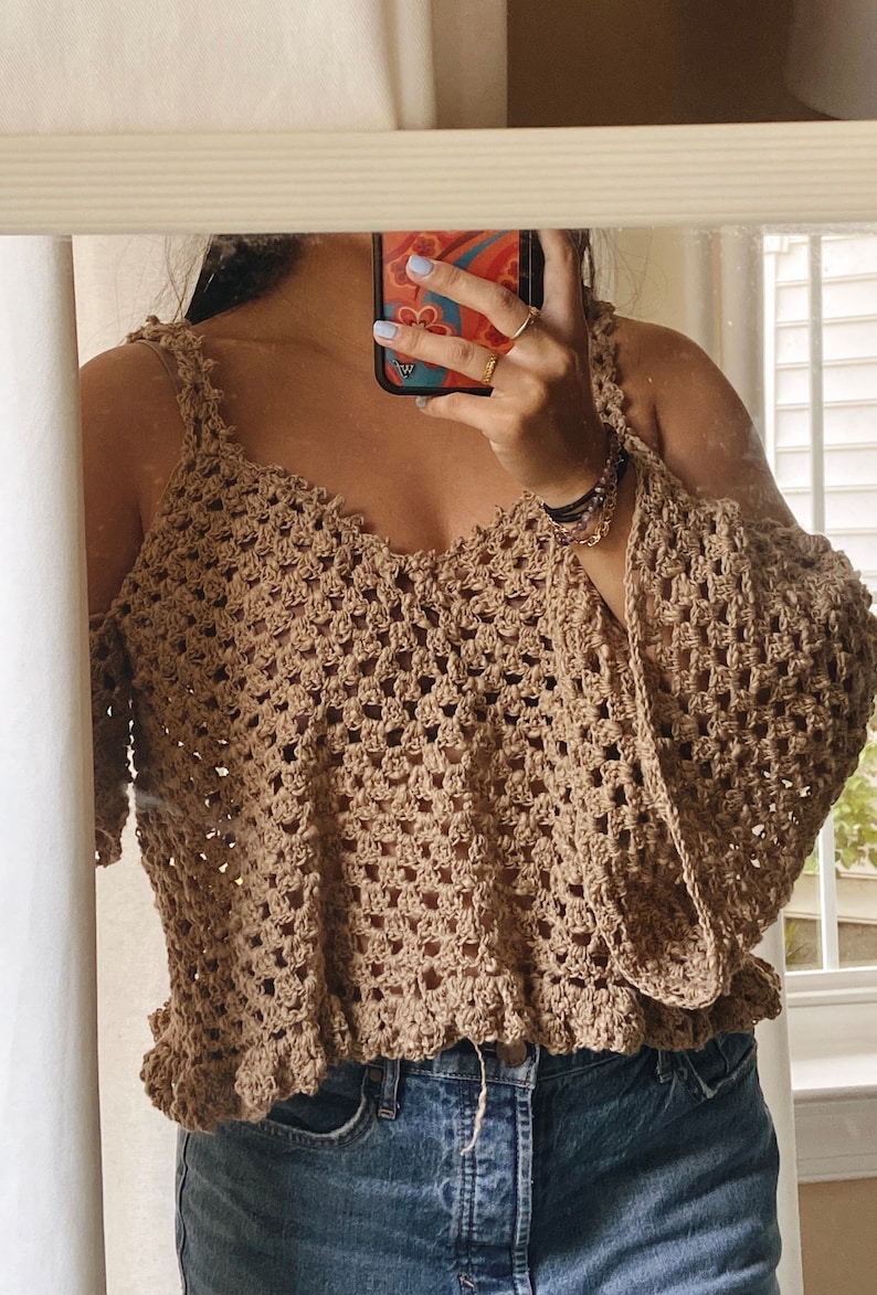 May include: A brown crochet top with a cold shoulder design and bell sleeves. The top has a crocheted trim around the neckline and hem. The top is worn over blue jeans.