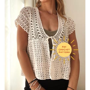 May include: Cream-colored, open-front crochet cardigan with short sleeves. The cardigan features a delicate, openwork pattern and a tie closure at the waist. A yellow circle with the text "PDF CROCHET PATTERN" is in the image.