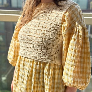 K&ouml;nnte beinhalten: Ein gelbes und wei&szlig;es Gingham-Kleid mit einem geh&auml;kelten Oberteil. Das Kleid hat einen runden Ausschnitt und Puff&auml;rmel.