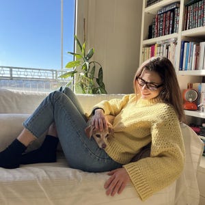 May include: A woman wearing a yellow crocheted sweater sits on a white couch with a small dog. The woman is wearing blue jeans and black socks. The dog is lying on the woman's lap. The woman is looking down at the dog.