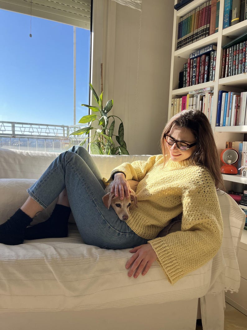 May include: A woman wearing a yellow crocheted sweater sits on a white couch with a small dog. The woman is wearing blue jeans and black socks. The dog is lying on the woman's lap. The woman is looking down at the dog.