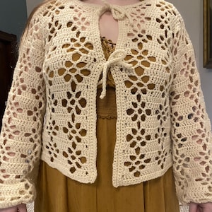 Pu&ograve; includere: Un cardigan all'uncinetto con un davanti a cravatta e un delicato motivo traforato. Il cardigan &egrave; di colore crema e ha maniche lunghe.