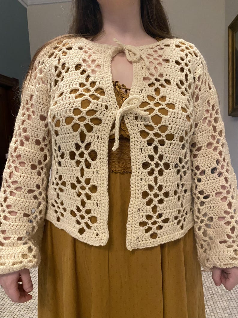 Pu&ograve; includere: Un cardigan all'uncinetto con un davanti a cravatta e un delicato motivo traforato. Il cardigan &egrave; di colore crema e ha maniche lunghe.
