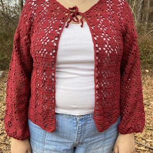 Pu&ograve; includere: Un cardigan di uncinetto rosso con un nodo sul collo. Il cardigan ha un motivo traforato e maniche lunghe.