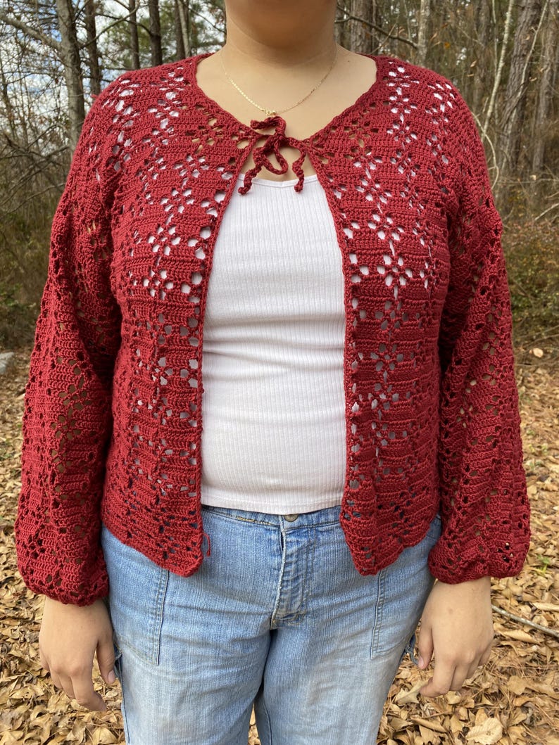 Pu&ograve; includere: Un cardigan di uncinetto rosso con un nodo sul collo. Il cardigan ha un motivo traforato e maniche lunghe.