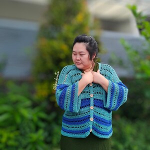 Op de afbeelding: Een turquoise en blauw gehaakt vest met klokmouwen en houten knopen. Het open ontwerp heeft horizontale strepen in verschillende tinten blauw en turquoise. Het vest wordt gedragen over een groene top en rok.