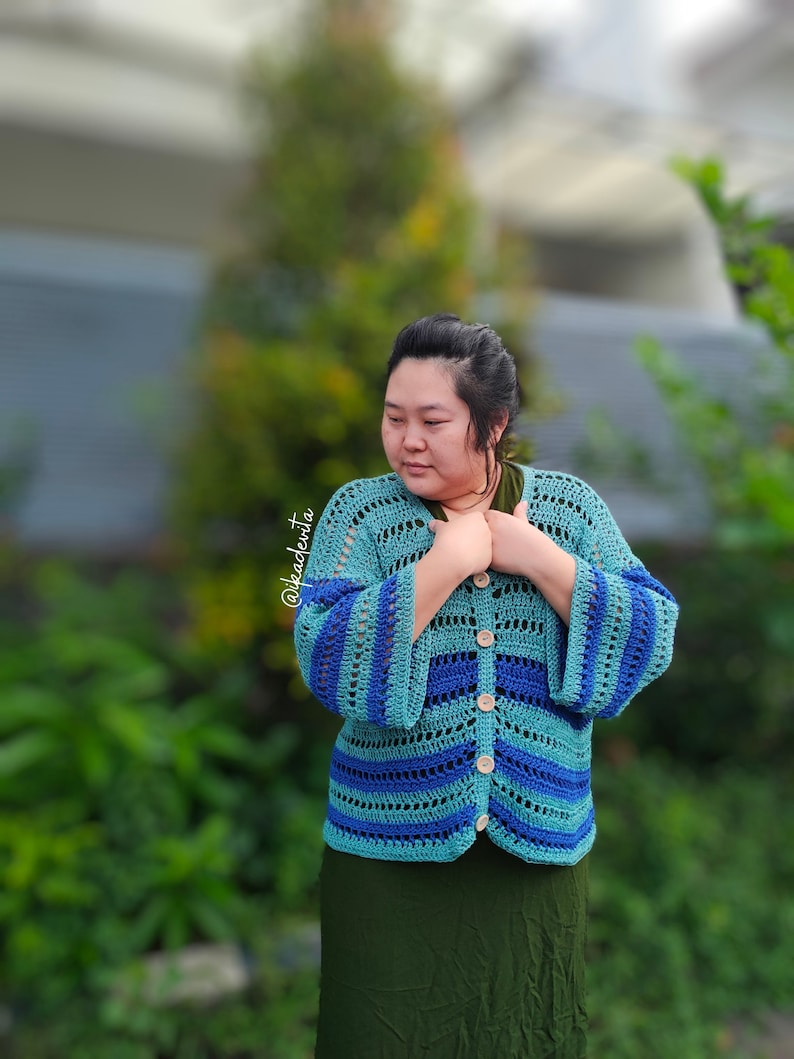 Op de afbeelding: Een turquoise en blauw gehaakt vest met klokmouwen en houten knopen. Het open ontwerp heeft horizontale strepen in verschillende tinten blauw en turquoise. Het vest wordt gedragen over een groene top en rok.