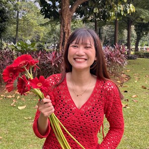 K&ouml;nnte beinhalten: Ein rotes Wickeloberteil aus H&auml;kelarbeit mit langen &Auml;rmeln und V-Ausschnitt. Das Oberteil wird mit einer blauen Jeans kombiniert. Die Person h&auml;lt einen Strau&szlig; roter Gerbera mit gr&uuml;nen Stielen. Der Hintergrund ist ein Park mit B&auml;umen und Gr&uuml;n.