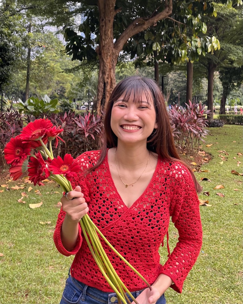 K&ouml;nnte beinhalten: Ein rotes Wickeloberteil aus H&auml;kelarbeit mit langen &Auml;rmeln und V-Ausschnitt. Das Oberteil wird mit einer blauen Jeans kombiniert. Die Person h&auml;lt einen Strau&szlig; roter Gerbera mit gr&uuml;nen Stielen. Der Hintergrund ist ein Park mit B&auml;umen und Gr&uuml;n.