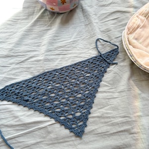 May include: A blue crocheted triangle scarf with a fringe edge. The scarf is lying on a white fabric surface.