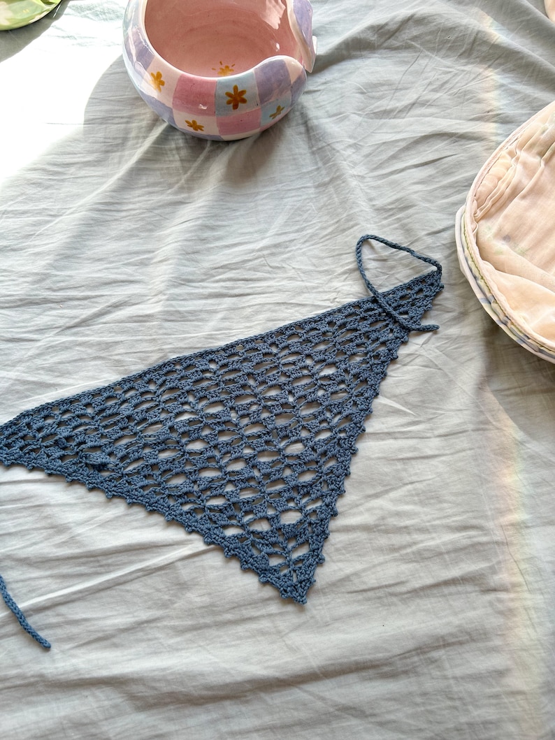 May include: A blue crocheted triangle scarf with a fringe edge. The scarf is lying on a white fabric surface.