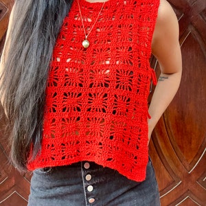 K&ouml;nnte beinhalten: Ein rotes H&auml;keltop mit quadratischem Muster und einem Band an der Seite. Das Top ist &auml;rmellos und hat einen runden Ausschnitt. Das Model tr&auml;gt das Top zu einer dunklen Jeans.