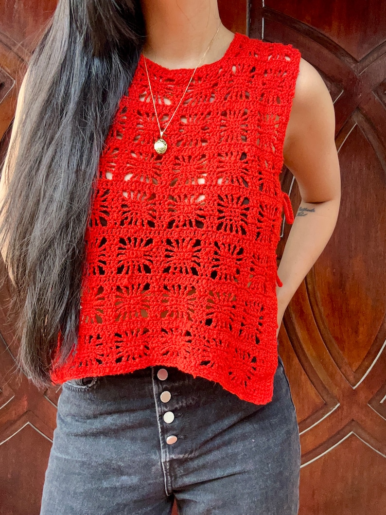 K&ouml;nnte beinhalten: Ein rotes H&auml;keltop mit quadratischem Muster und einem Band an der Seite. Das Top ist &auml;rmellos und hat einen runden Ausschnitt. Das Model tr&auml;gt das Top zu einer dunklen Jeans.