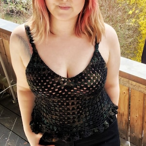 Op de afbeelding: Een vrouw draagt een zwart gehaakt topje met een delicate, kant-achtige rand. Het topje heeft een diepe V-hals en is gemaakt van zwart en groen garen. Ze draagt ook een zwarte jeans.