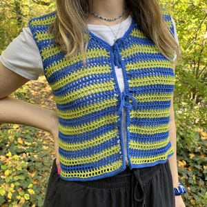 Op de afbeelding: Een blauw en geel gestreept gebreid vest met een strik sluiting. Het vest is mouwloos en kort.