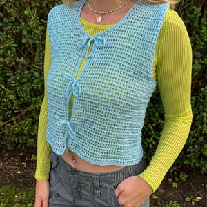 Op de afbeelding: Een lichtblauw gehaakt vest met een strik sluiting, gedragen over een limoengroen longsleeve shirt. Het vest heeft een losse, open structuur en een getailleerde lengte. Het model draagt grijze cargo broeken.