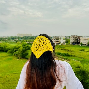 May include: A woman with long brown hair wearing a yellow crocheted headband. She is standing outdoors with a green field and buildings in the background.