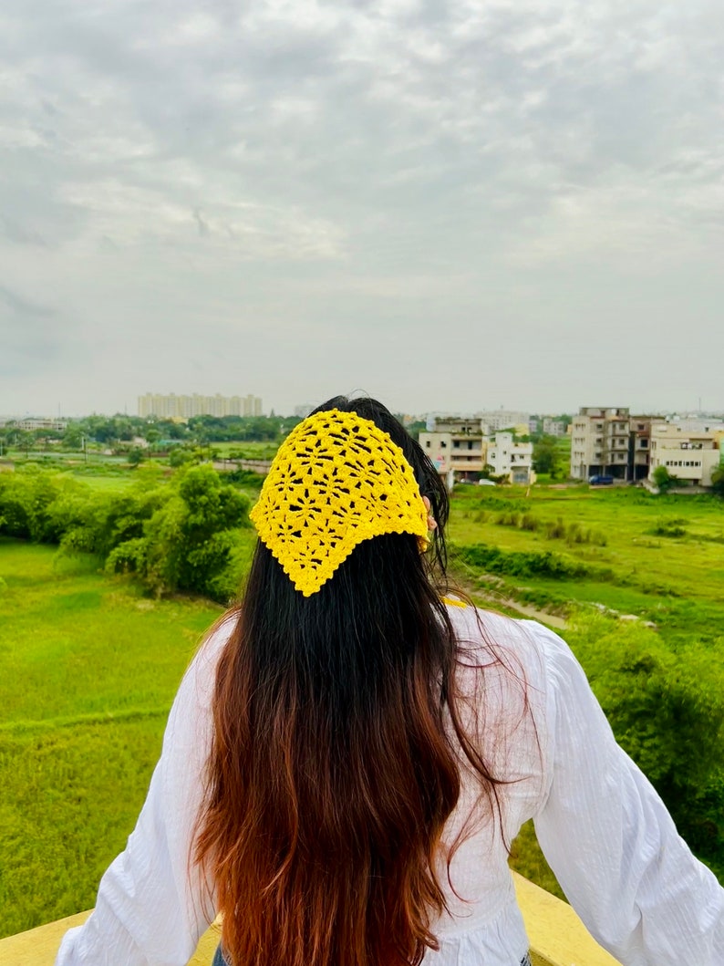 May include: A woman with long brown hair wearing a yellow crocheted headband. She is standing outdoors with a green field and buildings in the background.