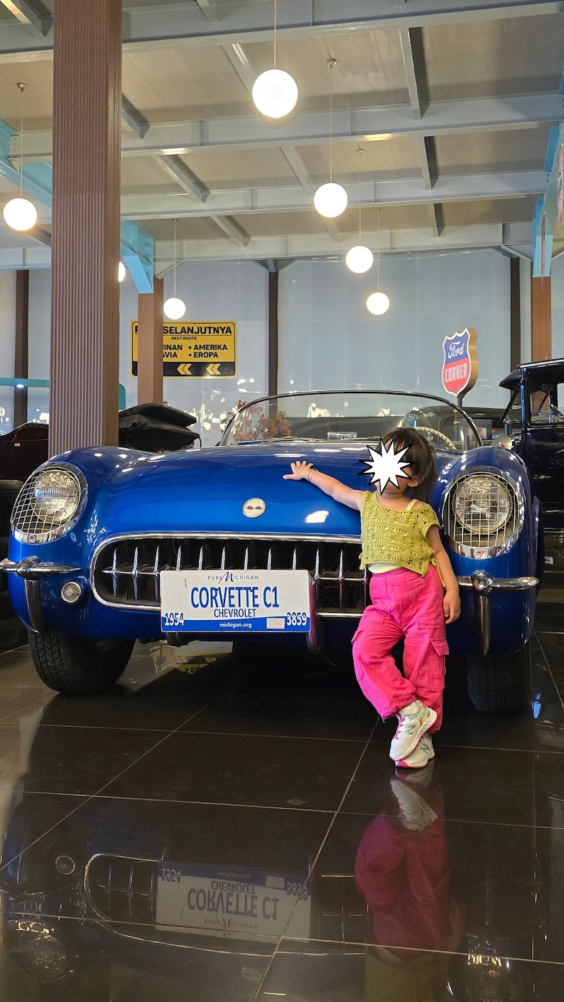 K&ouml;nnte beinhalten: Ein blauer Chevrolet Corvette C1 von 1954 mit einem Pure Michigan-Kennzeichen. Ein junges Kind steht neben dem Auto und lehnt sich an die Fahrert&uuml;r. Das Auto befindet sich in einem Museum mit einer wei&szlig;en Decke und Leuchten.