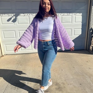 Op de afbeelding: Een lichtpaarse gehaakte cardigan met wijde mouwen. De cardigan wordt gedragen over een witte top, met een blauwe jeans en witte sneakers. De outfit is casual en comfortabel.