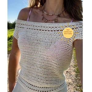 Op de afbeelding: Crèmekleurige, off-the-shoulder gehaakte top met korte mouwen. De top heeft een open weefpatroon. Een ketting wordt gedragen met de top, en een roze bandje is zichtbaar. De tekst "PDF Crochet Pattern" is inbegrepen.