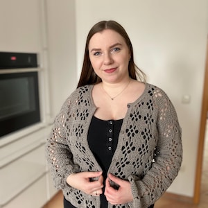 Pu&ograve; includere: Un cardigan di uncinetto grigio con chiusura a bottone. Il cardigan presenta un delicato motivo traforato ed &egrave; indossato sopra un top nero senza maniche e jeans neri.