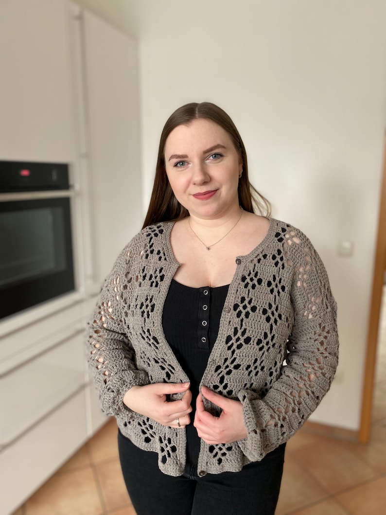 Pu&ograve; includere: Un cardigan di uncinetto grigio con chiusura a bottone. Il cardigan presenta un delicato motivo traforato ed &egrave; indossato sopra un top nero senza maniche e jeans neri.