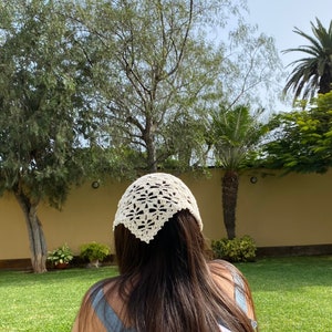 May include: A white crocheted bandana with a delicate floral pattern. The bandana is worn on the head of a person with long brown hair.