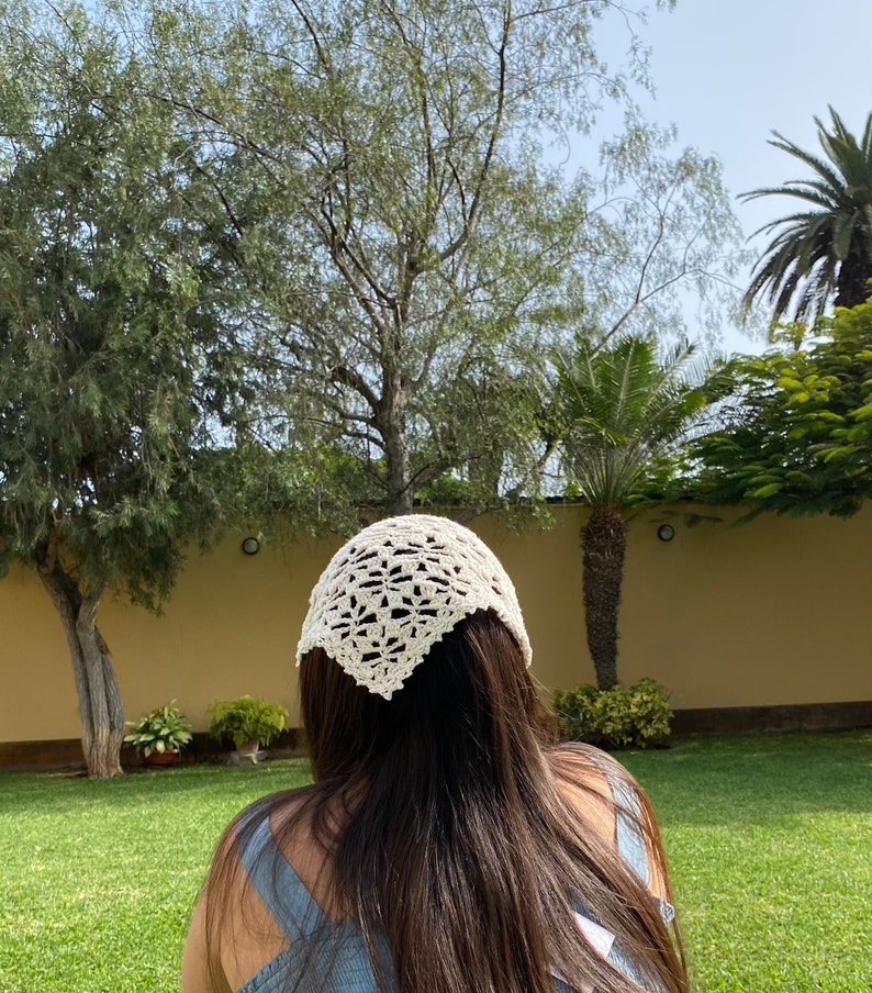 May include: A white crocheted bandana with a delicate floral pattern. The bandana is worn on the head of a person with long brown hair.