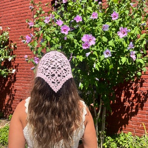 May include: A pink crocheted bandana tied around a person's head with long brown hair. The person is wearing a white crocheted top and is standing in front of a bush with purple flowers.