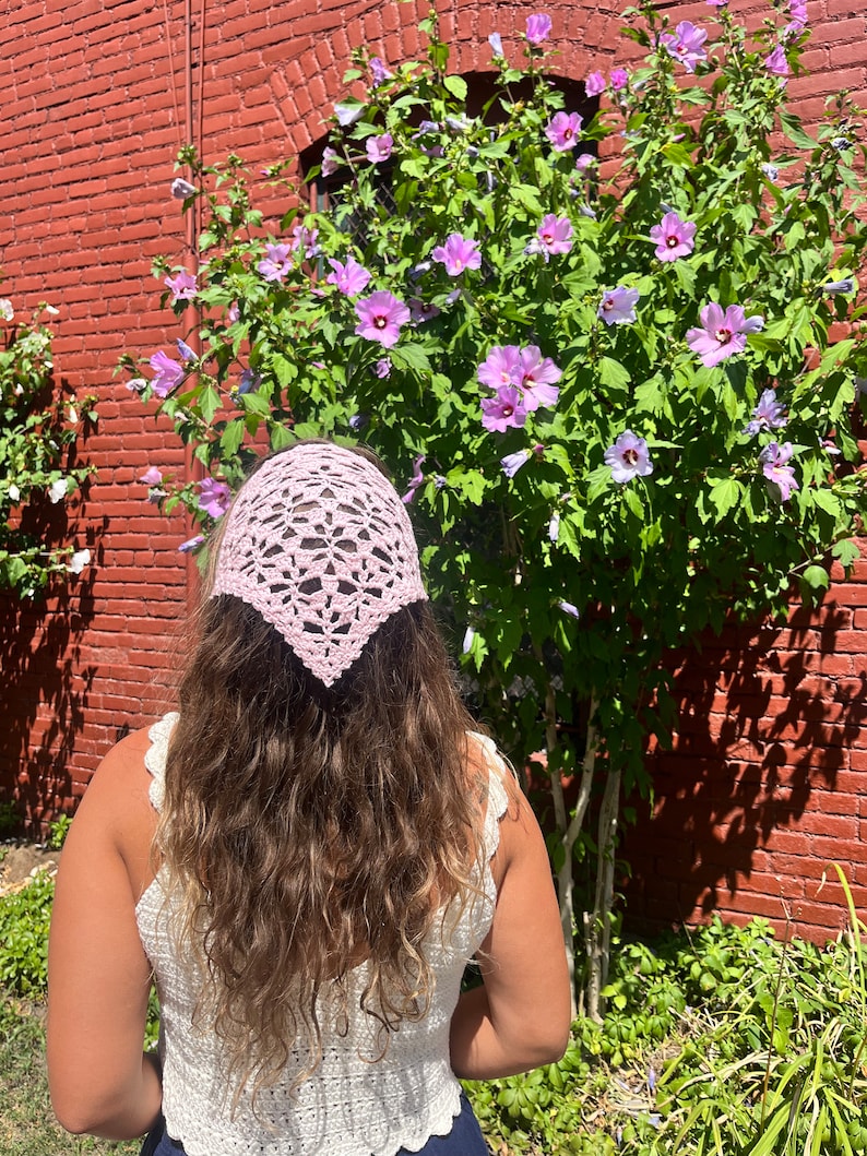 May include: A pink crocheted bandana tied around a person's head with long brown hair. The person is wearing a white crocheted top and is standing in front of a bush with purple flowers.