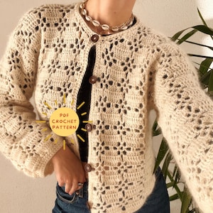 Könnte beinhalten: Eine gehäkelte Strickjacke mit Knopfleiste und einem zarten, durchbrochenen Muster. Die Strickjacke ist cremefarben und hat eine leicht flauschige Textur. Der Text "PDF CROCHET PATTERN" ist im Bild sichtbar.