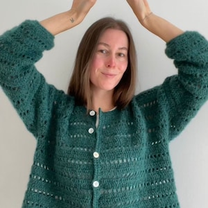 Op de afbeelding: Een teal groene cardigan met open weefsel en witte knopen. De lange mouwen, button-down trui is gemaakt van een zacht, getextureerd garen. Het model draagt een blauwe jeans. De cardigan is een handgemaakt, gezellig kledingstuk.