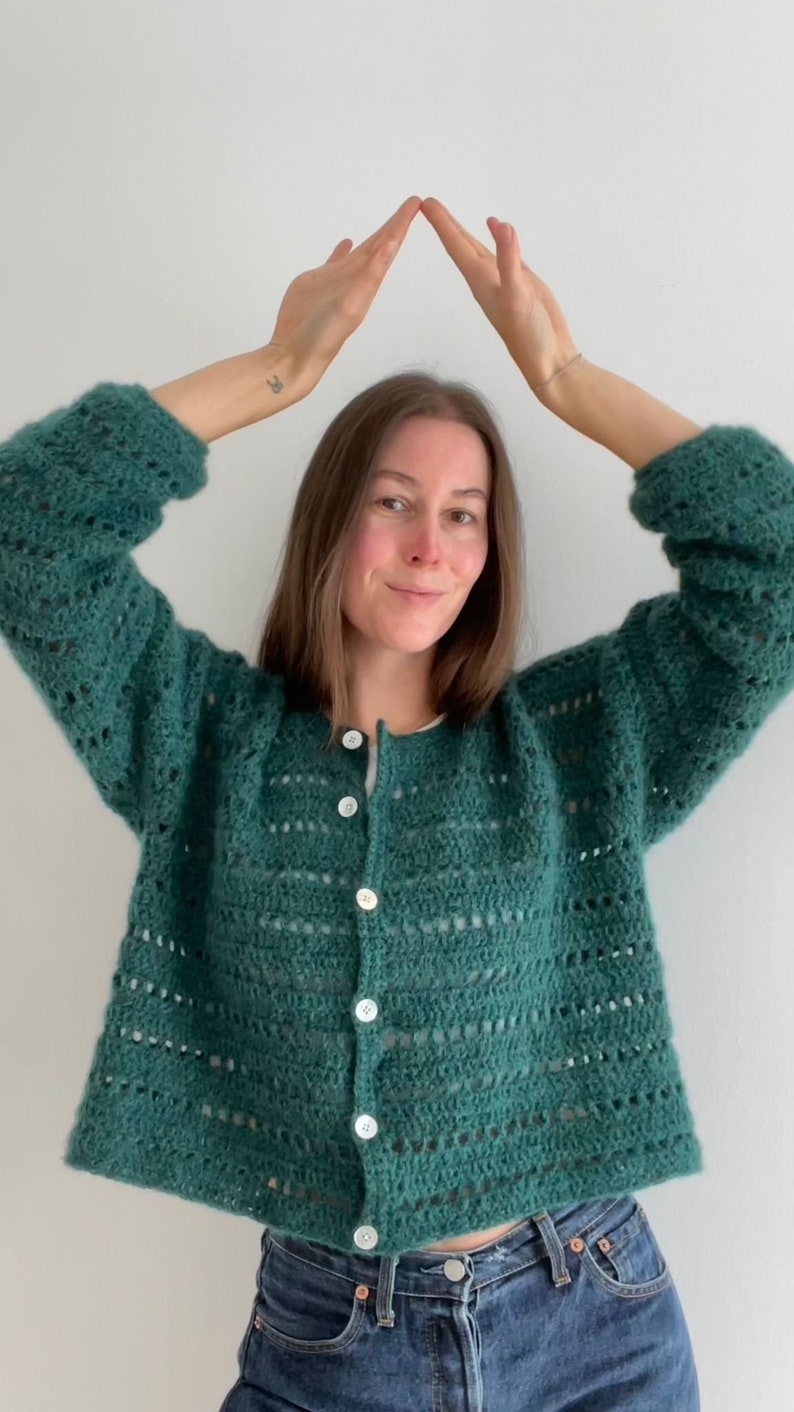 Op de afbeelding: Een teal groene cardigan met open weefsel en witte knopen. De lange mouwen, button-down trui is gemaakt van een zacht, getextureerd garen. Het model draagt een blauwe jeans. De cardigan is een handgemaakt, gezellig kledingstuk.