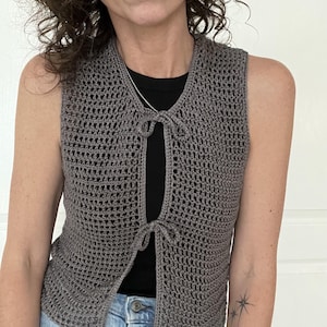 Op de afbeelding: Een grijs gebreid vest met een strik aan de voorkant. Het vest is mouwloos en heeft een losse pasvorm.