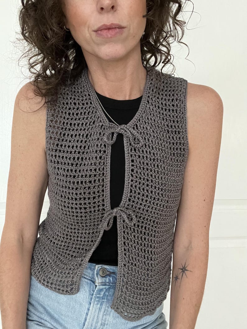 Op de afbeelding: Een grijs gebreid vest met een strik aan de voorkant. Het vest is mouwloos en heeft een losse pasvorm.