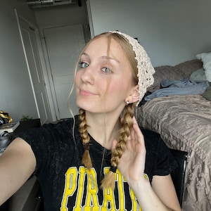 May include: A woman with blonde hair in two braids wearing a white crocheted headband and a black t-shirt with the word "PIRATE" printed in yellow.