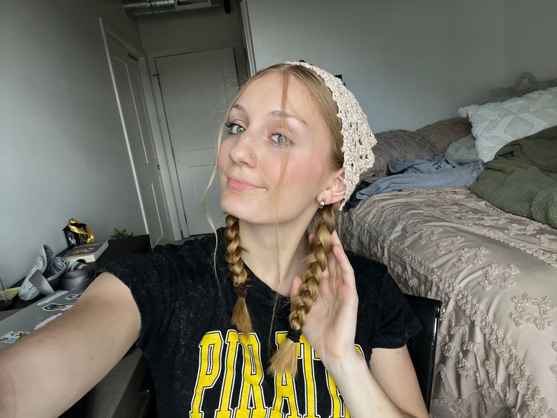 May include: A woman with blonde hair in two braids wearing a white crocheted headband and a black t-shirt with the word "PIRATE" printed in yellow.