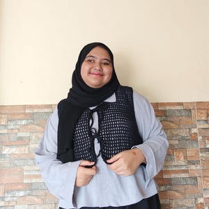 Op de afbeelding: Een vrouw draagt een lichtblauw overhemd met knopen en daaroverheen een zwarte gehaakte vest. Ze draagt ook een zwarte hoofddoek en een zwarte broek.