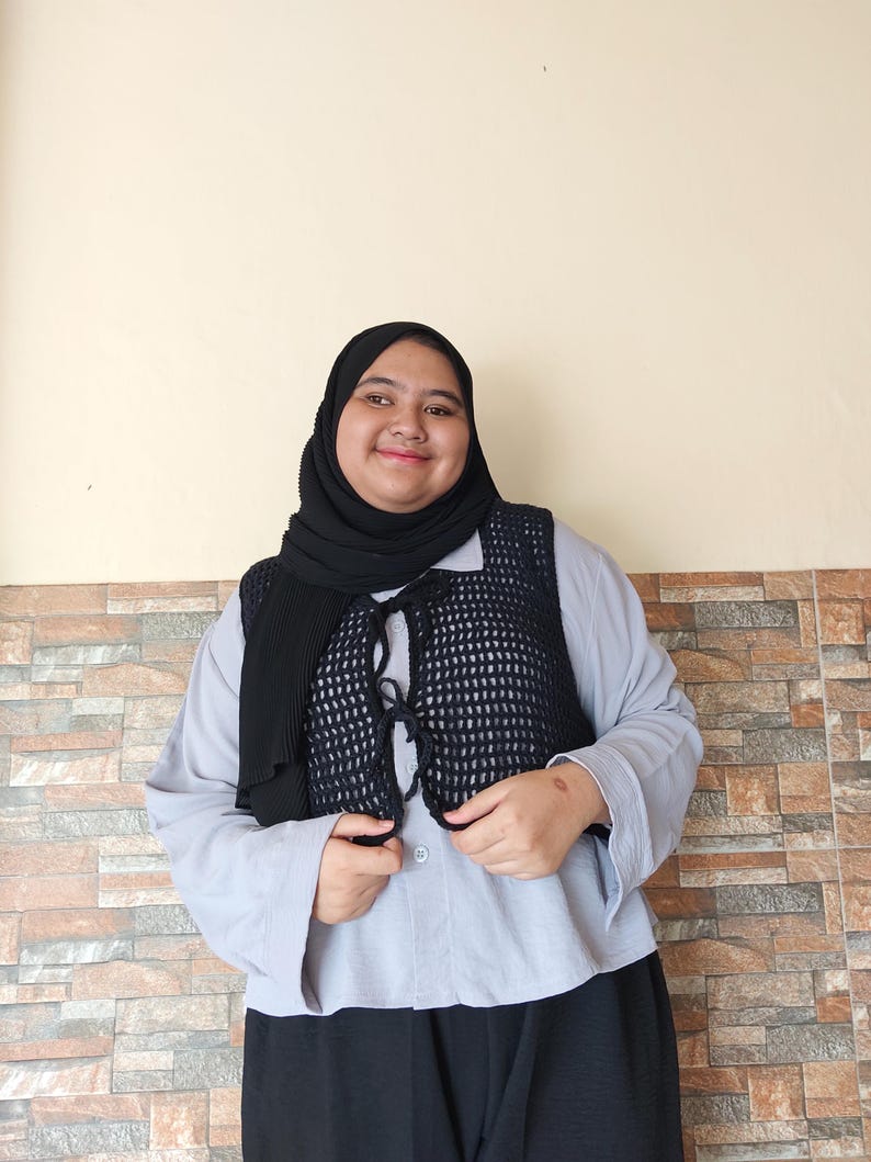 Op de afbeelding: Een vrouw draagt een lichtblauw overhemd met knopen en daaroverheen een zwarte gehaakte vest. Ze draagt ook een zwarte hoofddoek en een zwarte broek.