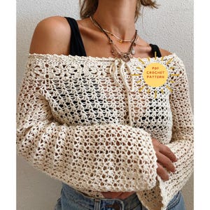 Op de afbeelding: Crèmekleurige gehaakte top met lange mouwen en een strik aan de halslijn. De top heeft een open, luchtig ontwerp en wordt off-shoulder gedragen. De afbeelding bevat een gele cirkel met de tekst "PDF CROCHET PATTERN".