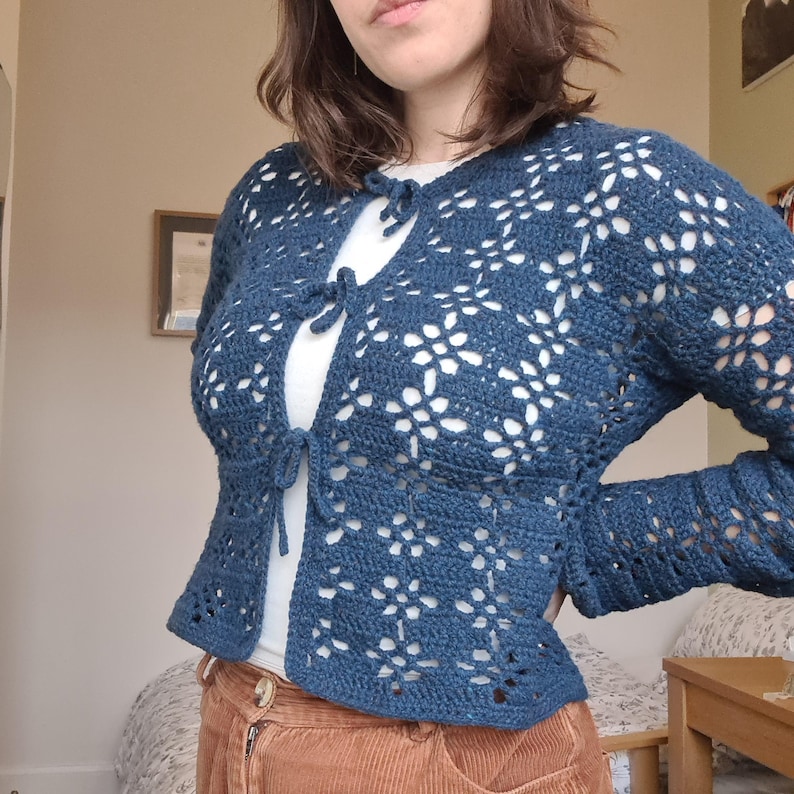 Pu&ograve; includere: Un cardigan di uncinetto blu con un davanti a cravatta e un motivo floreale.